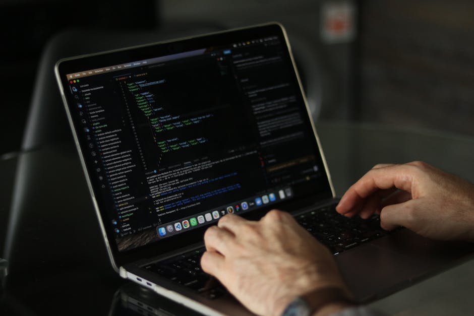 A person's hands typing on a closed silver laptop with a black keyboard, positioned on a dark table. The laptop screen displays a code editor with lines of multicolored programming code on a dark back